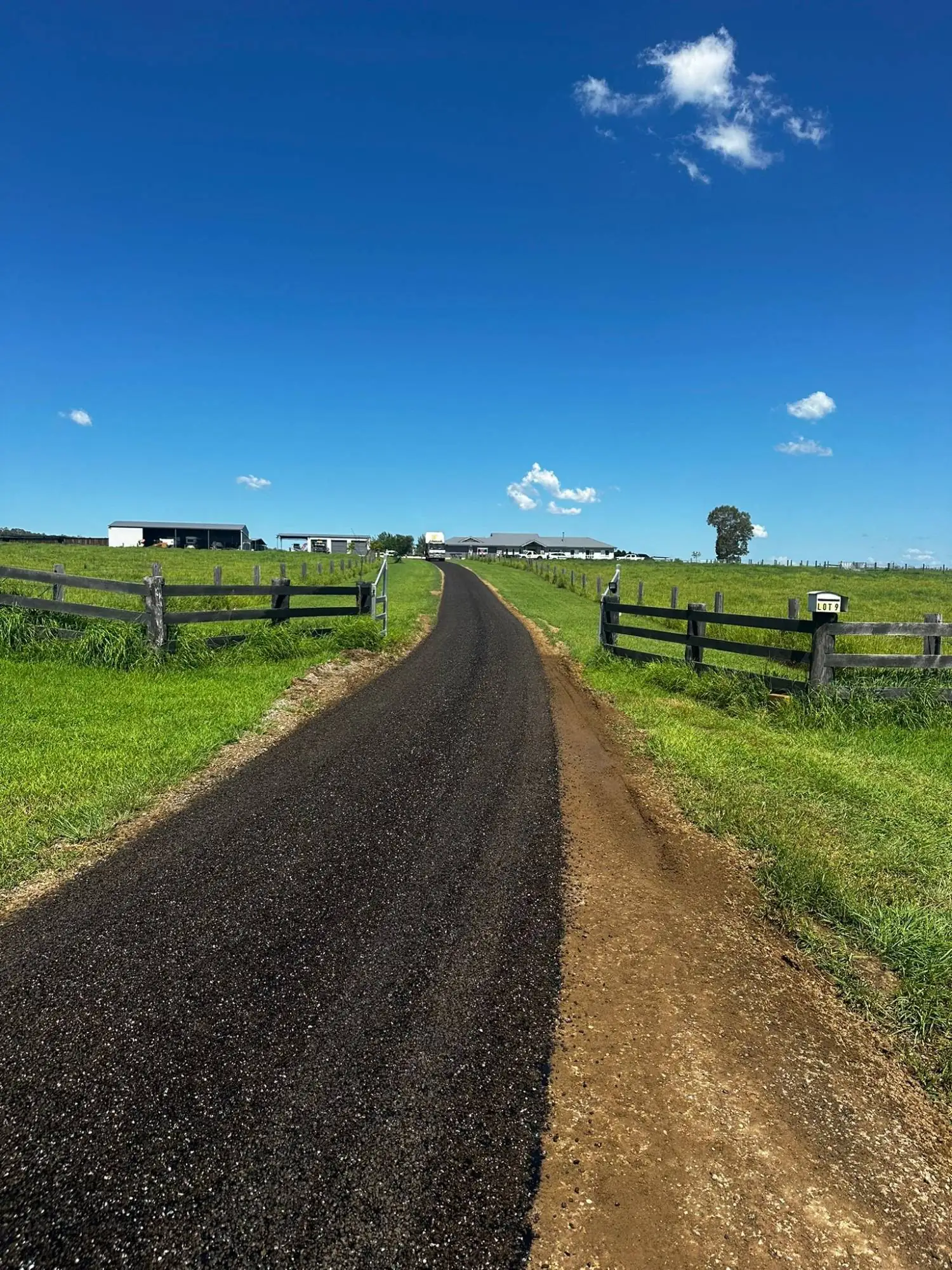 A freshly paved rural driveway leads through green farmland toward farm buildings